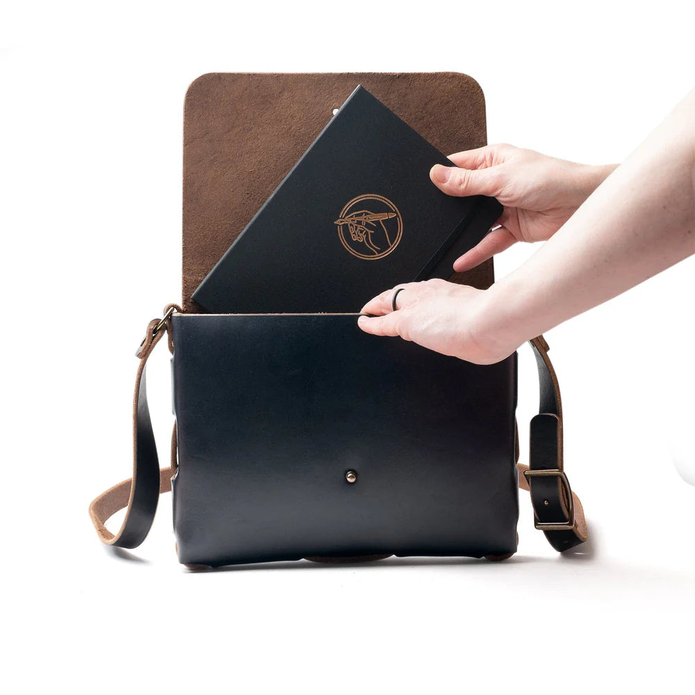 Person placing a black leather item into a brown leather bag on a white background