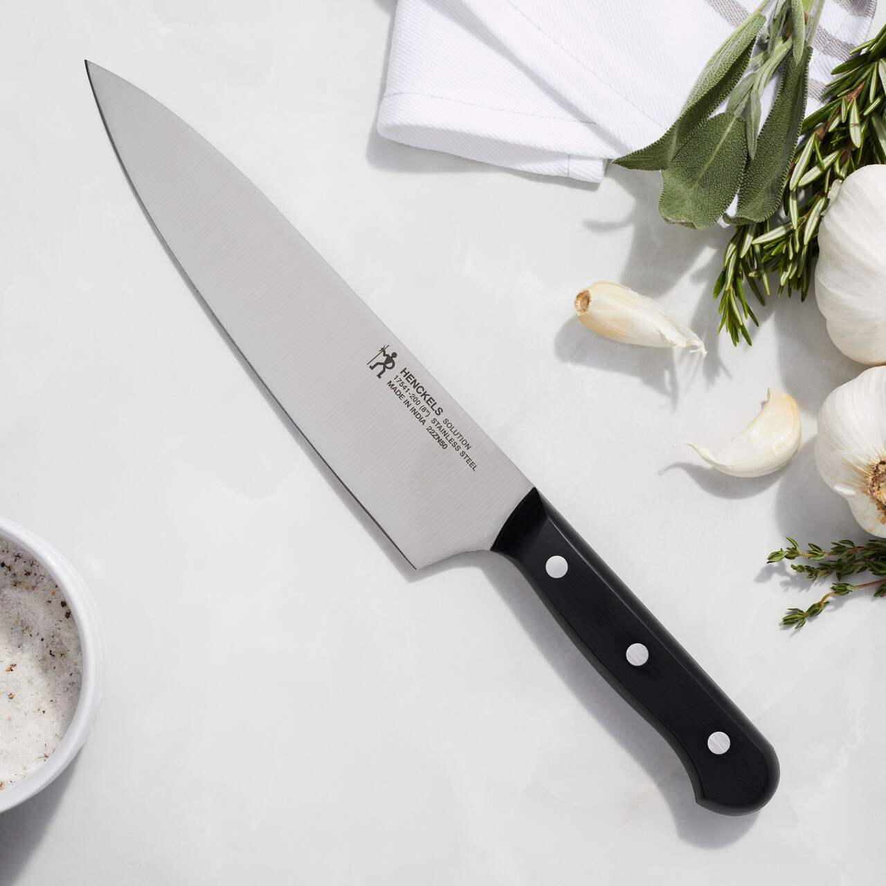 Chef's knife with black handle on a white surface with herbs and garlic